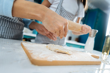 Good cooks. Loving cute little daughter making some dough and holding a rolling pin while her mom helping her and they wearing aprons