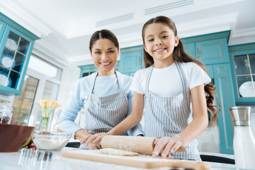 Fototapeta premium We feel great. Pretty alert dark-haired schoolgirl and mother smiling and making some dough for cookies while wearing aprons and using a rolling pin