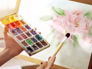 Obraz premium Artist painting on easel in studio. Authentic body part hands of girl paints with brush and set palette watercolor paints . Drawing of spring flowers.