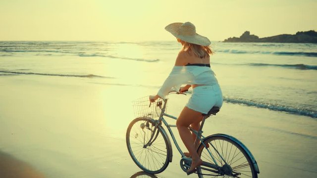 Woman in white and hat mooving on her bicycle along beach sand at summer time under burning sunset sky, lifestyle carefree