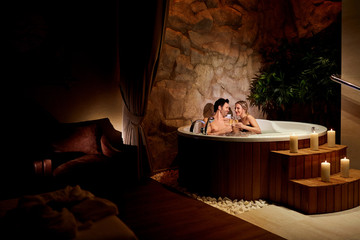 A loving couple with a glass of champagne in a jacuzzi with candles relax. - obrazy, fototapety, plakaty