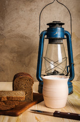 Kerosene lantern, cutting board, clay pot with milk, rye bread and a knife on a wooden table. Still-life in a rustic style