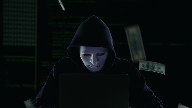 Hacker in mask working on laptop, dollar bills falling down, money laundering