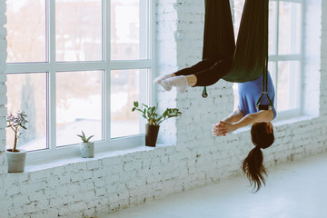 Young woman doing fly yoga and stretches in front of window on white background in loft interior studio.