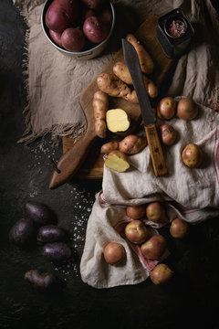 Variety of raw uncooked organic potatoes different kind and colors red, yellow, purple on wooden cutting board with kitchen towels over black table. Top view. Dark rustic style