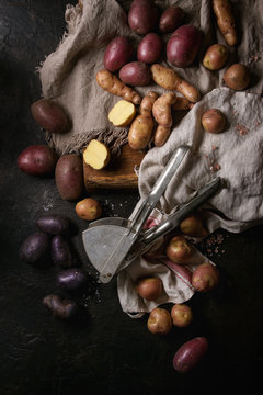 Variety of raw uncooked organic potatoes different kind and colors red, yellow, purple on wooden cutting board with kitchen towels over black table. Top view. Dark rustic style. Toned image