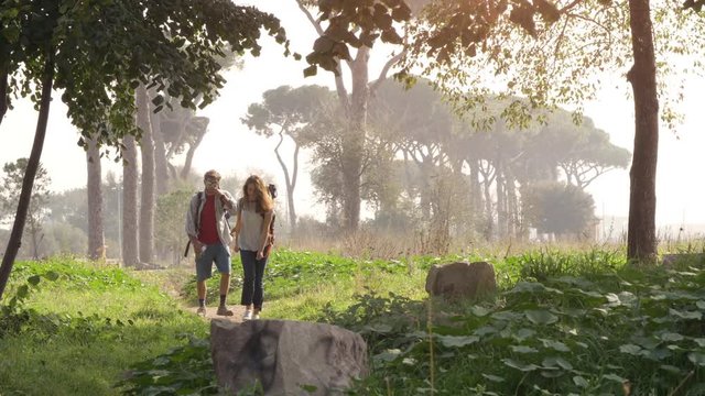Young happy couple backpackers tourists walking in the nature along dirt road in aqueduct park ruins in rome countryside on misty morning with guitar and sleeping bag slow motion