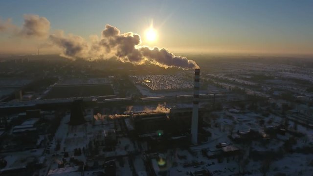 A bird`s eye view of a sky high chimney with a white smoke flux at a splendid sunset in winter. The cityscape is urban and good