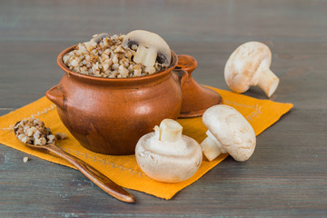 Clay pot with hot buckwheat porridge and mushrooms champignons on a gray wooden table. The concept of a healthy diet, vegetarian food. Selective focus.