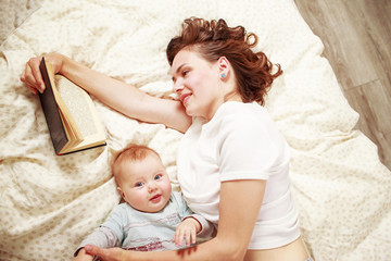 young smiling woman reading a book while lying on bed with baby