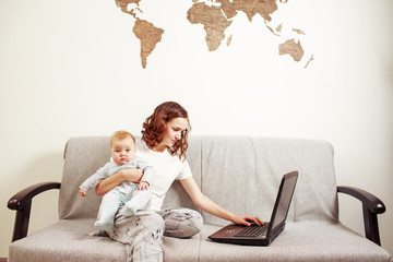 Young woman in white t-shirt and grey pants holding a baby while working using laptop