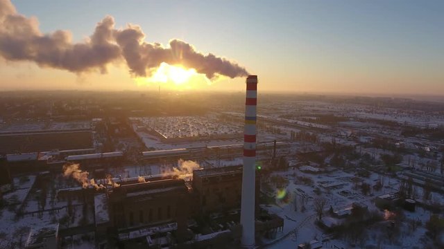 A bird`s eye view of a sky-high chimney with a slow flux of smoke at a wonderful sunset in winter. It stands near a plant. and houses.