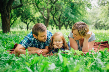 Fototapeta premium Young happy family of three lying on blanket in the park having fun. Happy parenting concept. Little girl with mother and father outdoors