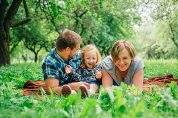 Fototapeta premium Young happy family of three lying on blanket in the park having fun. Happy parenting concept. Little girl with mother and father outdoors