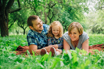 Fototapeta premium Young happy family of three lying on blanket in the park having fun. Happy parenting concept. Little girl with mother and father outdoors