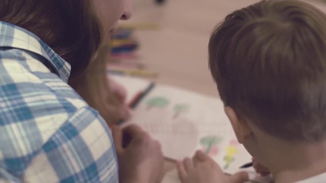 Rear View - Positive Caring Woman Helps Her Little Charming Son To Draw A Drawing With The Colored Pencils Sitting At The Table At Home