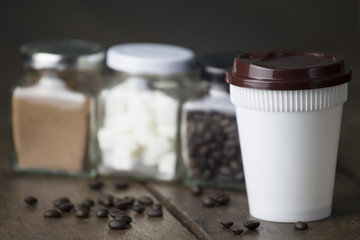 Hot coffee white plastic cup with brown cap and three glass jar on old wooden table with low light.