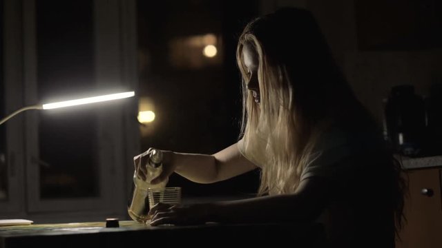 Young woman pouring alcohol in glass from a bottle and drinking in dark room