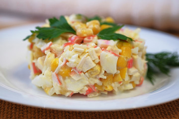 On a white plate salad of crab sticks, corn, boiled rice, eggs and with the addition of mayonnaise. The dish is decorated with leaves of green parsley. Close up view.