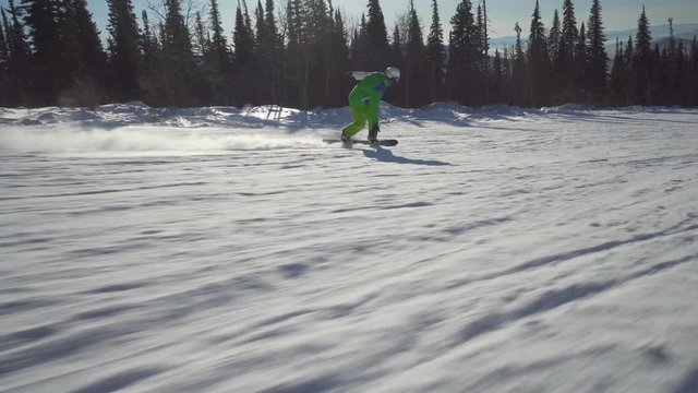 Sportsman in bright green clothes is riding on board upon a slope of the hill at sunny winter day