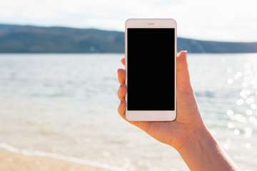 A woman is holding a phone against a sea beach. Blank screen
