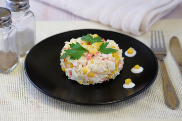 On a black plate salad of crab sticks, corn, boiled rice, eggs and with the addition of mayonnaise. The dish is decorated with leaves of green parsley. There are cutlery nearby.
