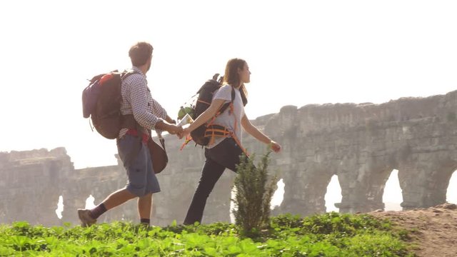 Young lovely couple backpackers tourists walking holding hands toward roman aqueduct arches in parco degli acquedotti park ruins in rome on romantic misty sunrise with guitar and sleeping bag slow