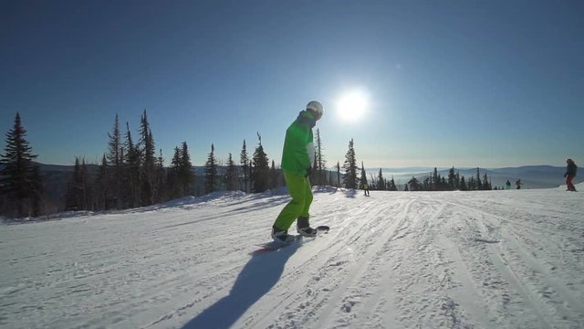 Sportsman in bright green clothes is jumping on snowboard upon a slope of the hill at winter sunny day