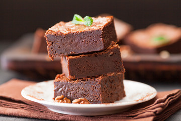 Chocolate brownie square pieces in stack on white plate with walnuts, decorated with mint leaves and cocoa on black background. Delicious dessert. Dark mood. Close up photography. Selective focus