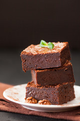 Chocolate brownie square pieces in stack on white plate with walnuts, decorated with mint leaves and cocoa on black background. Delicious dessert. Dark mood. Close up photography. Selective focus