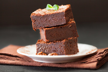 Chocolate brownie square pieces in stack on white plate with walnuts, decorated with mint leaves and cocoa on black background. Delicious dessert. Dark mood. Close up photography. Selective focus
