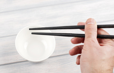 Hand holding Chinese chopsticks next to white bowl on wooden table.
