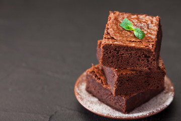 Fototapeta na wymiar Chocolate brownie square pieces in stack on white plate with walnuts, decorated with mint leaves and cocoa on black background. Delicious dessert. Dark mood. Close up photography. Selective focus