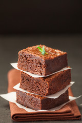 Chocolate brownie square pieces in stack on white plate with walnuts, decorated with mint leaves and cocoa on black background. Delicious dessert. Dark mood. Close up photography. Selective focus