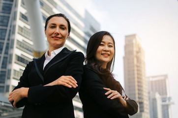 two smiling business asian and caucasian woman in formally dressed looking into camera with her arms crossed standing in modern city, confident, team leader, successful and entrepreneurship concept