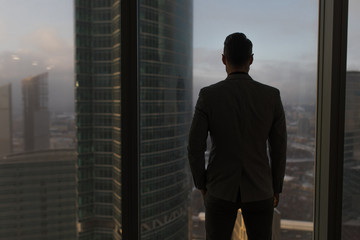 Rearview of a businessman looking out the window at the city. - obrazy, fototapety, plakaty