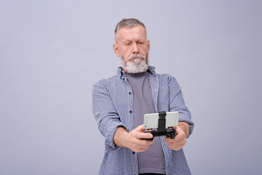 Emotional Senior Man With Video Game Controller For Smartphone On Grey Background