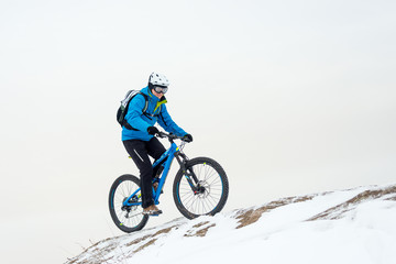 Fototapeta na wymiar Cyclist in Blue Riding Mountain Bike on Rocky Winter Hill Covered with Snow. Extreme Sport and Enduro Biking Concept.