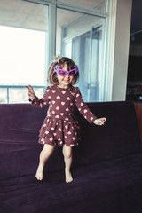 Portrait of cute adorable little girl with funny glasses jumping dancing on couch indoors. Preschooler having fun at home. Lifestyle happy childhood concept. Kid celebrating holiday