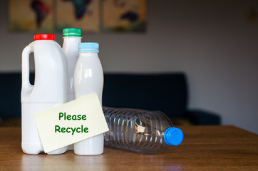 Plastic bottles with paper sticker with phrase `Please recycle` standing on the wooden table at home in the daylight with dark blurred map of planet Earth on the background