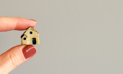 Closeup of tiny plastic house holding in the woman's fingers on blurred grey wall background