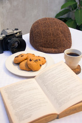 Fototapeta na wymiar Table with chocolate chips cookies, book, cup of coofee, camera and hat