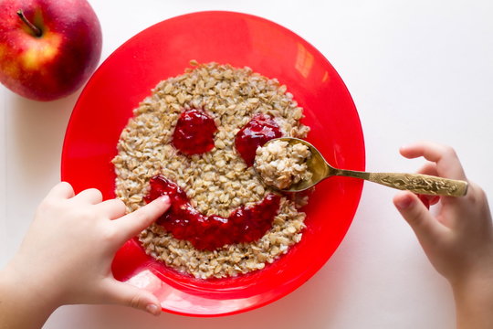A child eats a Breakfast of porridge oats out of a red dish, try a finger painted jam,smile