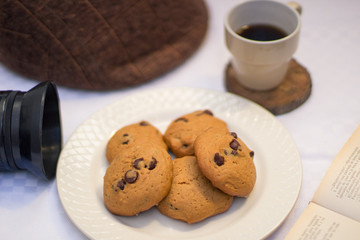 Table with chocolate chips cookies, book, cup of coofee, camera and hat