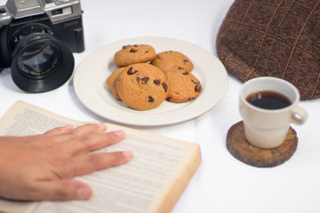 Table with chocolate chips cookies, book, cup of coofee, camera and hat