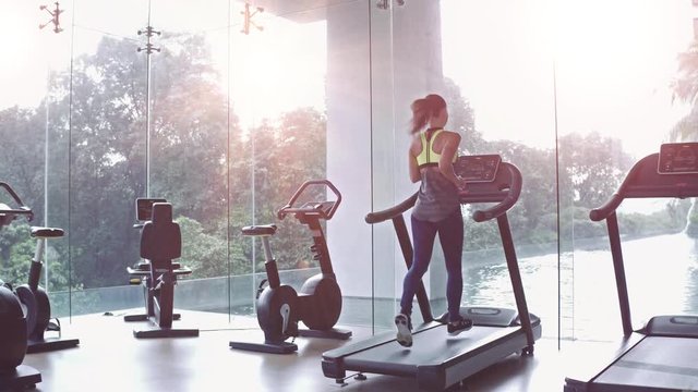 Woman press button and start running on treadmill at a panoramic window and listening to music via headphone at gym. Concept of healthy lifestyle.