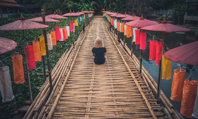 A girl in a black T-shirt is sitting with her back to the camera on a bamboo path decorated with bright Thai umbrellas. Vacation in Thailand. Chiang Mai