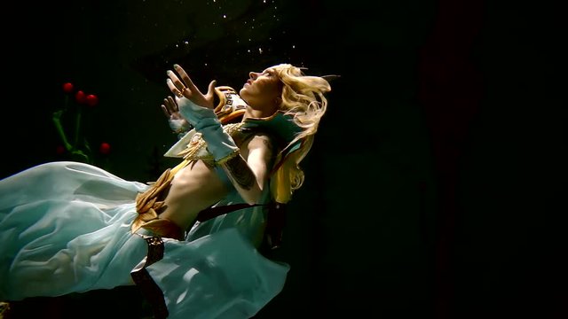 Divine woman with blonde hairs wearing elf costume is swimming on her back underwater