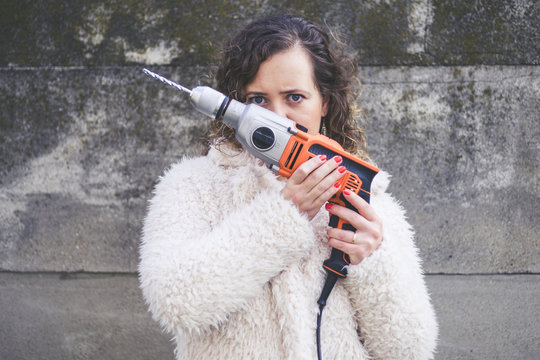Beautiful curly woman wearing fur coat and posing a drill in front of a wall. International Womans Day concept.
