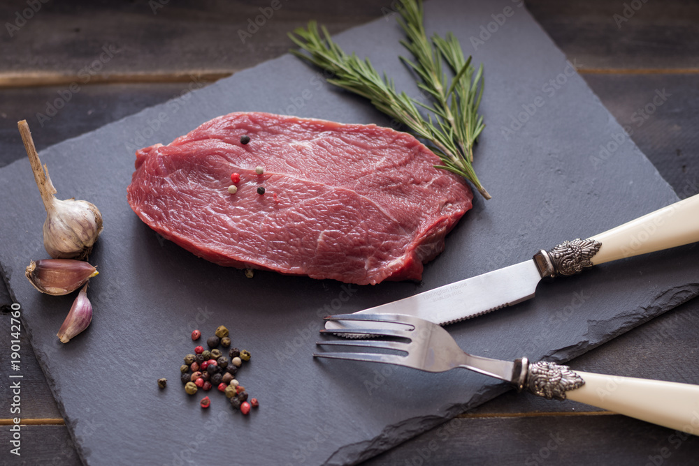 Wall mural Raw beef steak with rosemary, peppers garlik on dark stone, fork and knife